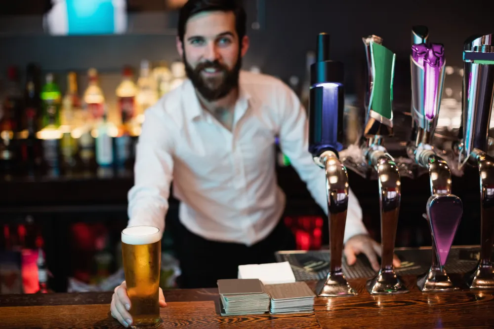A smiling bartender with a beard serves a beer in a lively Texas bar, showcasing good customer service and a positive work environment.