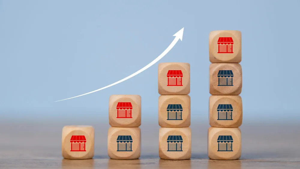 A bar graph made of wooden blocks with storefront icons, showing a white arrow pointing up, symbolizing business expansion.